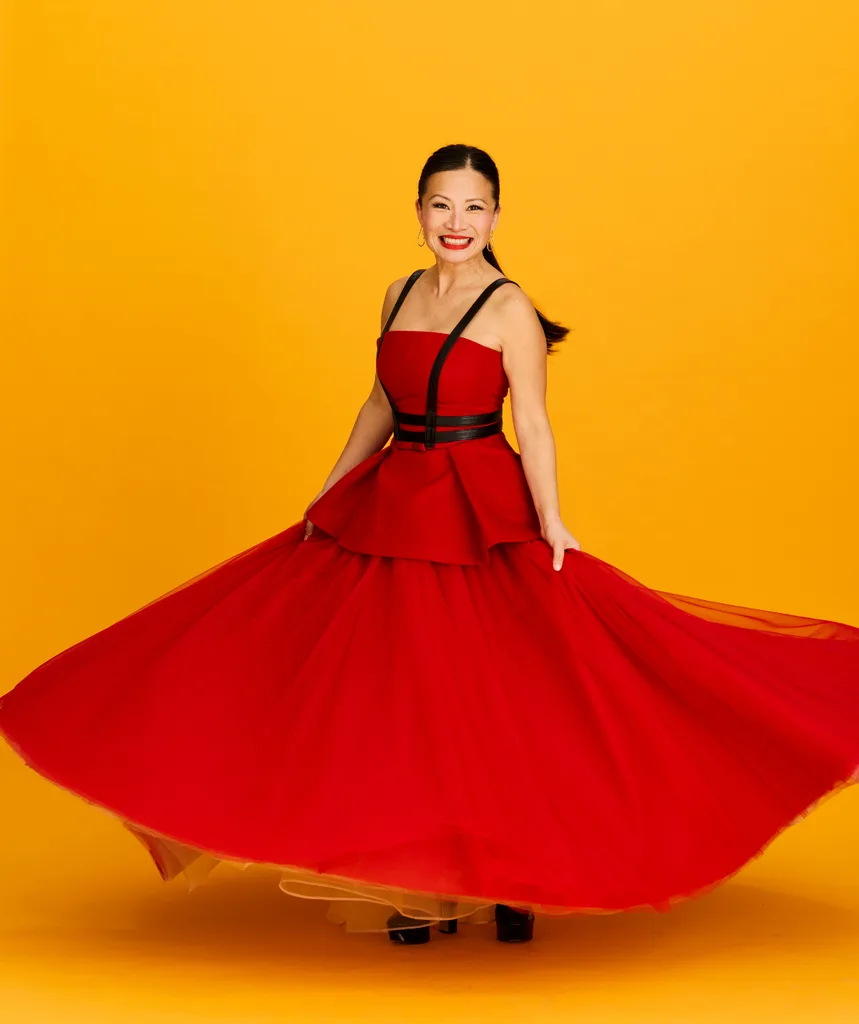 Poh, in a floor-length red dress, poses in front of an orange background.