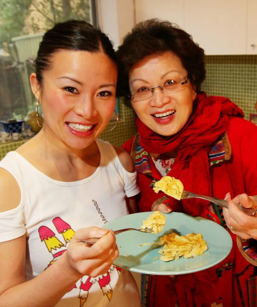 Poh and her mother smile at the camera as they eat food from a plate.