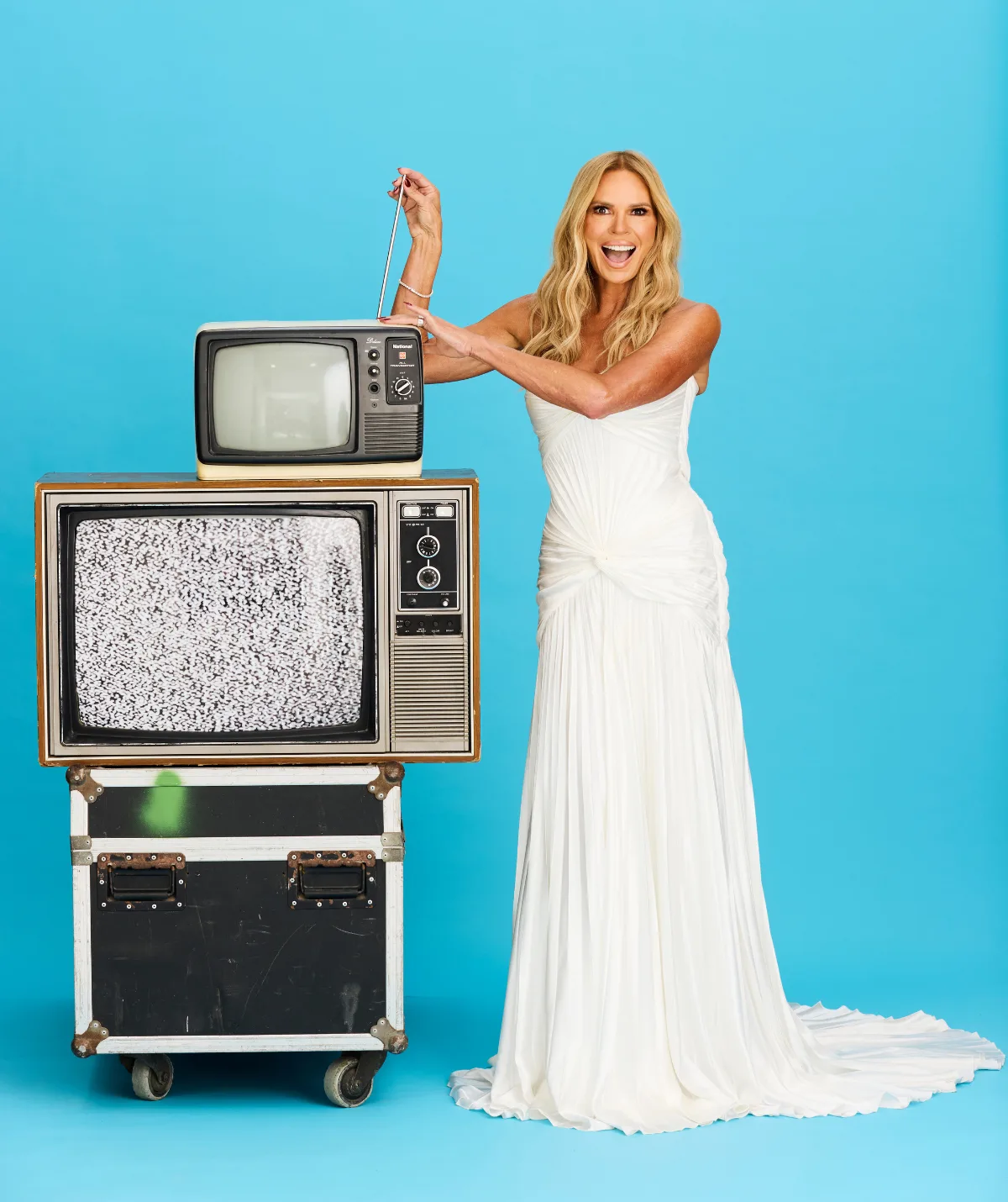 Sonia plays with the antennae on top of a tower of old TV's and sound equipment. She is smiling in a white fitted sleeveless gown. Blonde hair is out and waved. She is against a bright blue backdrop.