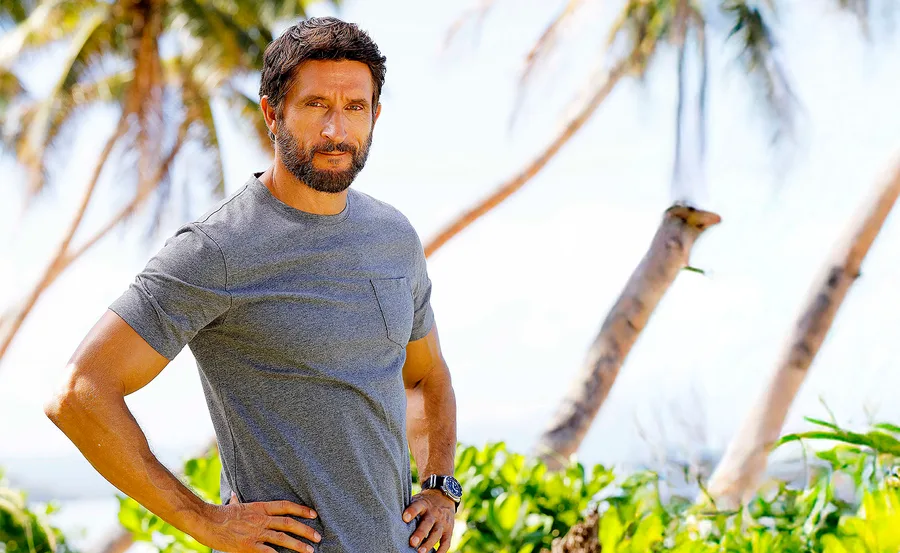 Jonathan LaPaglia, in a blue tshirt, stands with his hands on hips in front of palm trees.