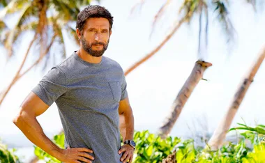 Jonathan LaPaglia, in a blue tshirt, stands with his hands on hips in front of palm trees.