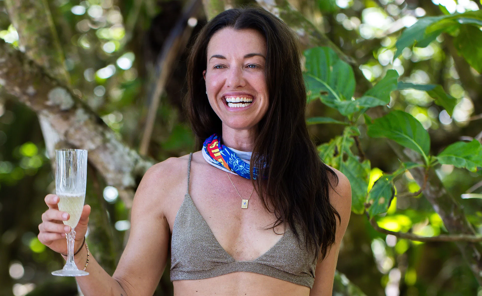 Parvati Shallow, wearing a bikini and with a Survivor buff around her neck, poses in front of vegetation, smiling.