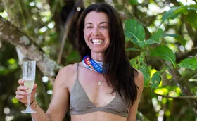 Parvati Shallow, wearing a bikini and with a Survivor buff around her neck, poses in front of vegetation, smiling.