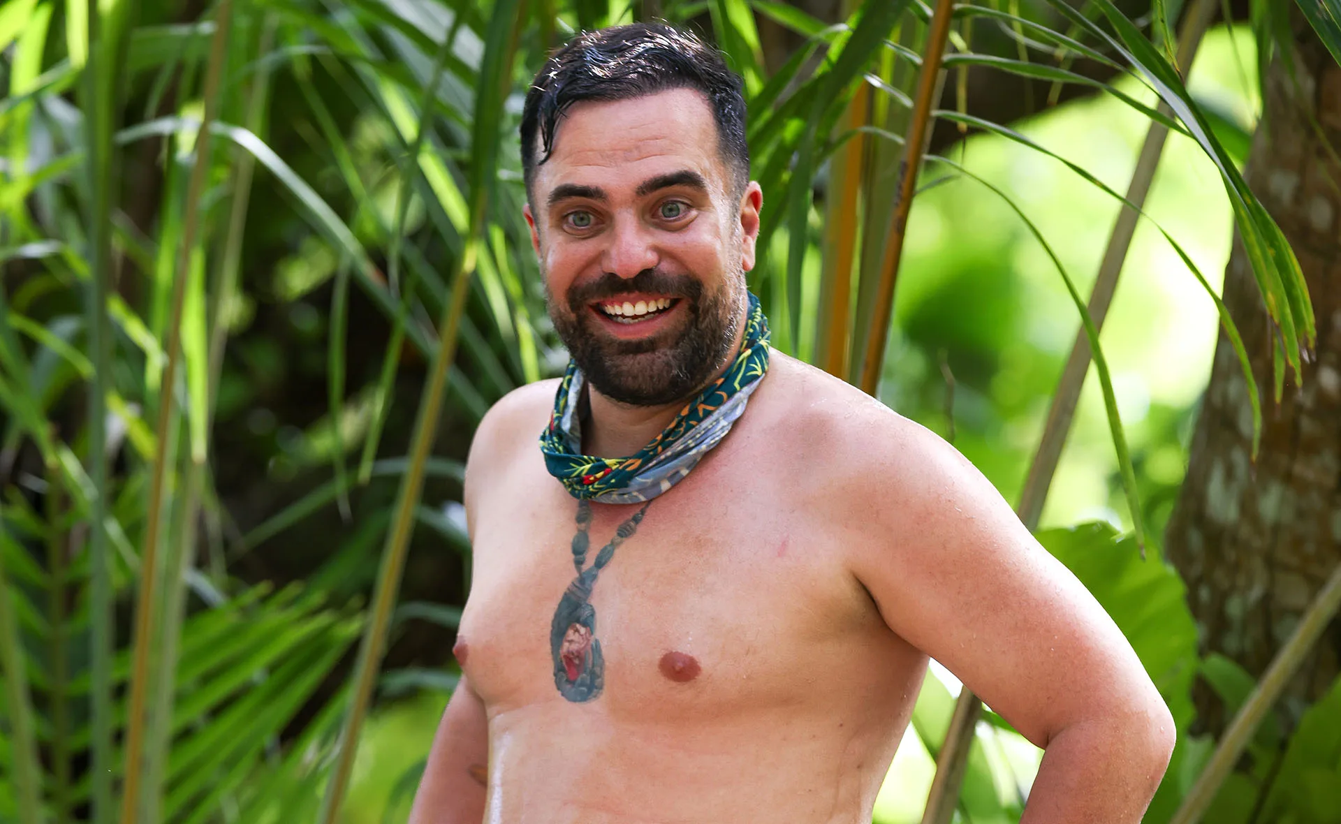 George, shirtless, wearing his Survivor buff and showing off his immunity idol tattoo, smiles at the camera.