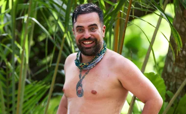 George, shirtless, wearing his Survivor buff and showing off his immunity idol tattoo, smiles at the camera.