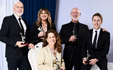 Executive producer Vincent Sheehan, stars Julia Zemiro and Glenn Butcher, and director Tom Peterson stand in a group around star and creator Kitty Flanagan, who is sitting on a chair, with a blue curtain behind them. All are holding Logies.