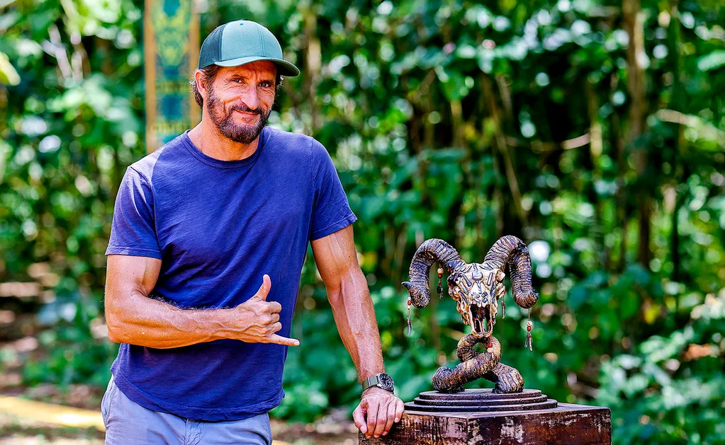 Jonathan LaPaglia, wearing a blue tshirt and a cap, smiles at the camera while standing in front of trees on the set of Survivor.