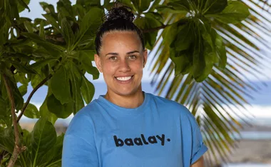 Kirby, wearing a blue t-shirt and smiling, stands in front of tropical vegetation