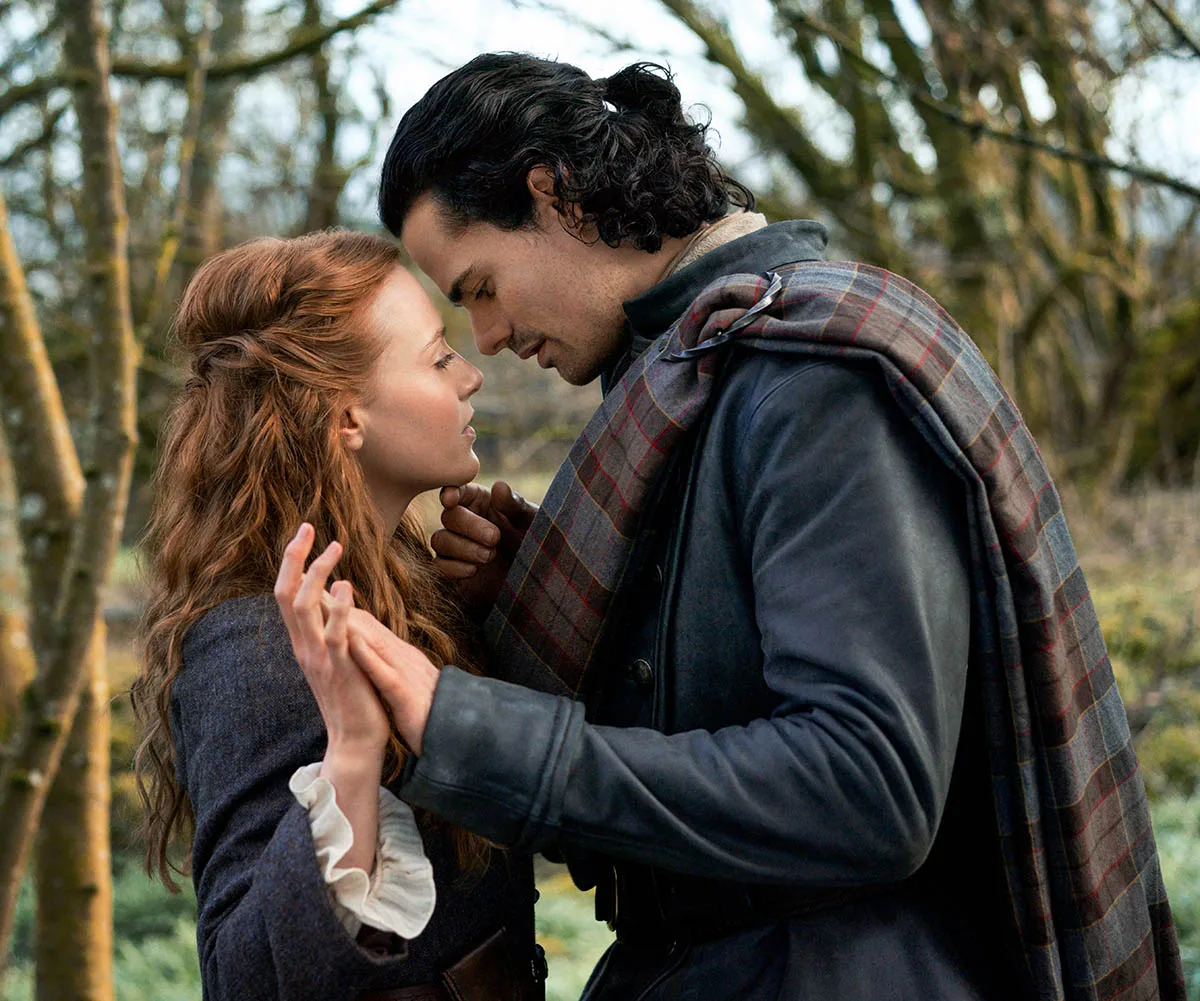 Harriet and Jamie, in character on the set of Blood Of My Blood, hold hands, Jamie's forehead resting on Harriet's.