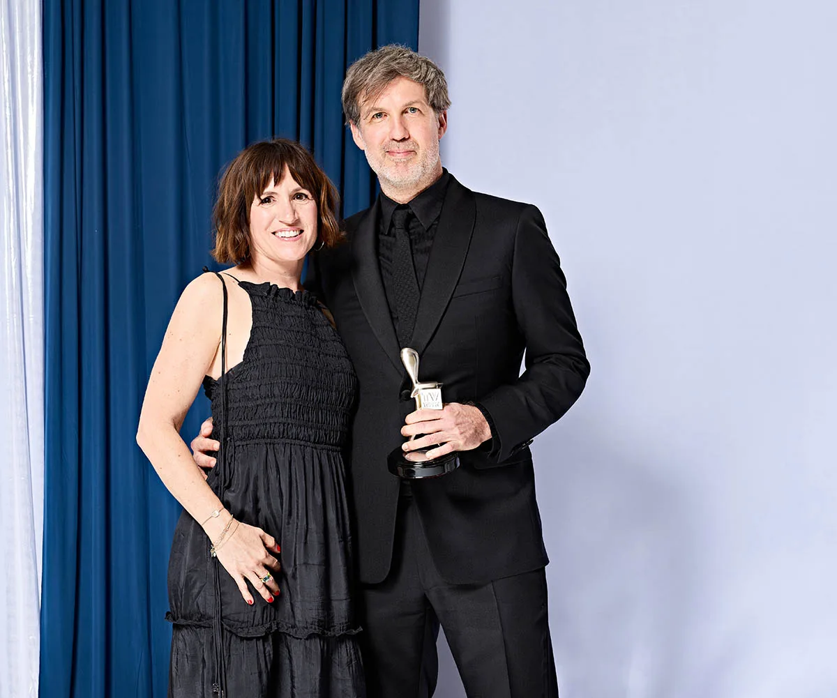 Joe Brumm holds the Logie, with his arm around his wife Suzy, with a blue curtain behind them, backstage at the Logies,