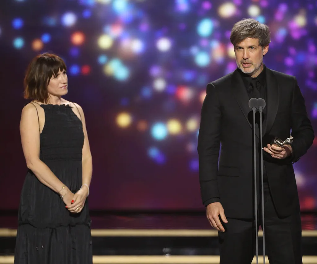 Joe Brumm stands on the Logies stage behind a microphone, while his wife Suzy stands next to him, looking at him..