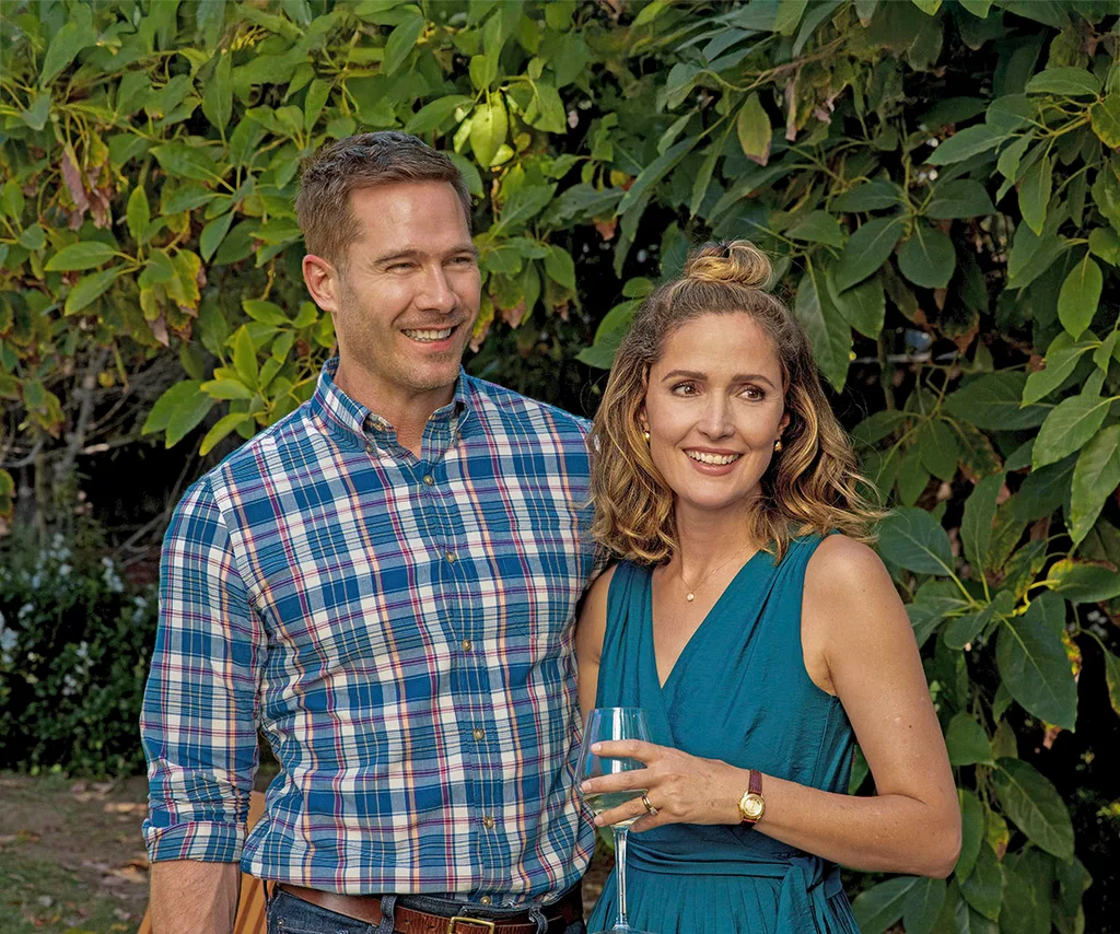 Luke MacFarlane as Charlie, Sylvia's husband. She holds a glass of champagne and the pair smile while holding each other in front of greenery on season two of Platonic.