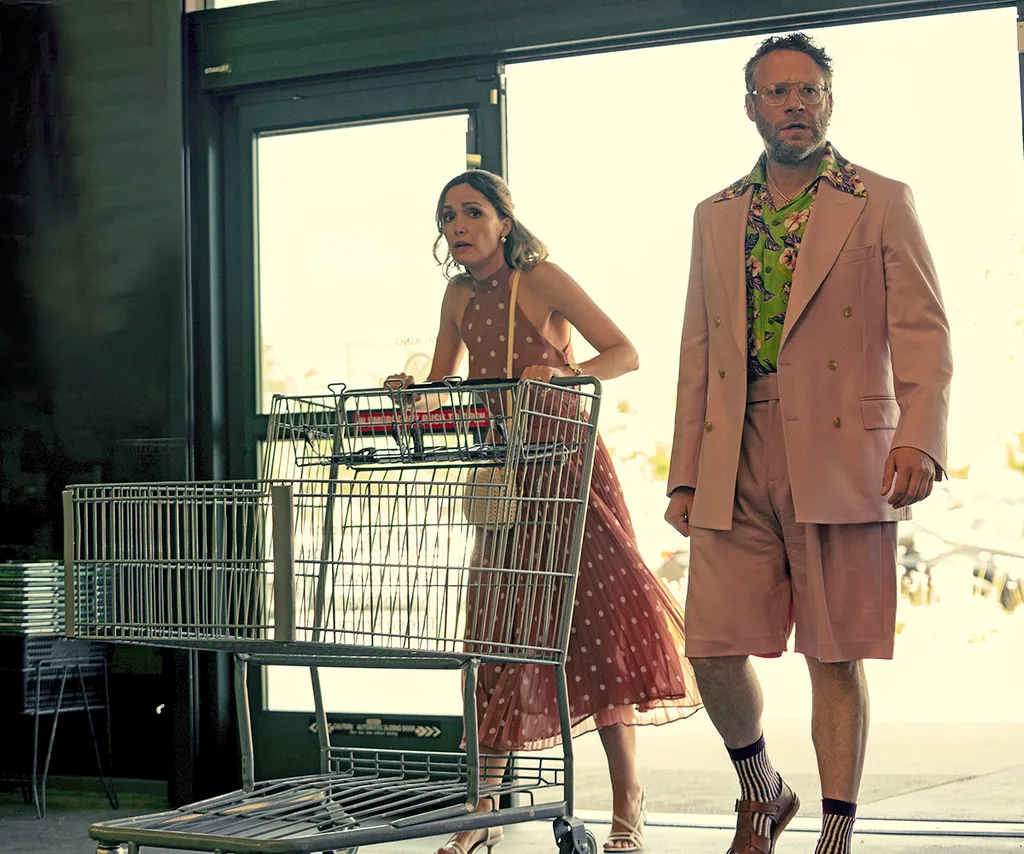 Rose Byrne and Seth Rogan as Sylvia and Will on set of Platonic season two. Sylvia pushing a trolley into a liquor store next to Will in a pink suit jacket and shorts.