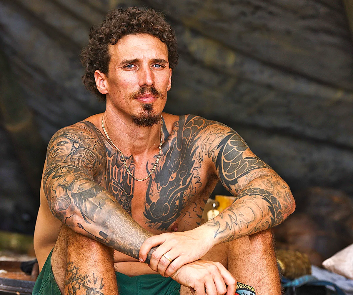 Luke, shirtless and tattooed, sits with his arms resting on his knees, looking thoughtful. 