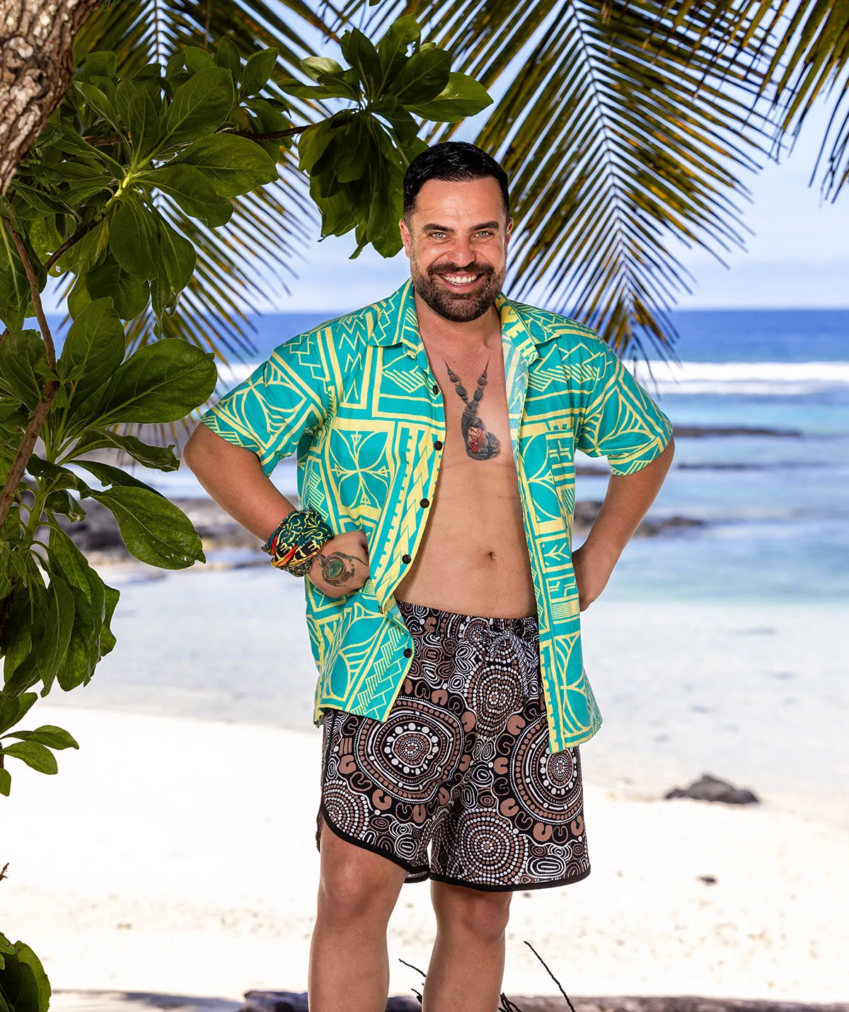 George, wearing a colourful shirt and showing off his immunity idol tattoo, stands with his hands on his hips on a beach in Samoa.