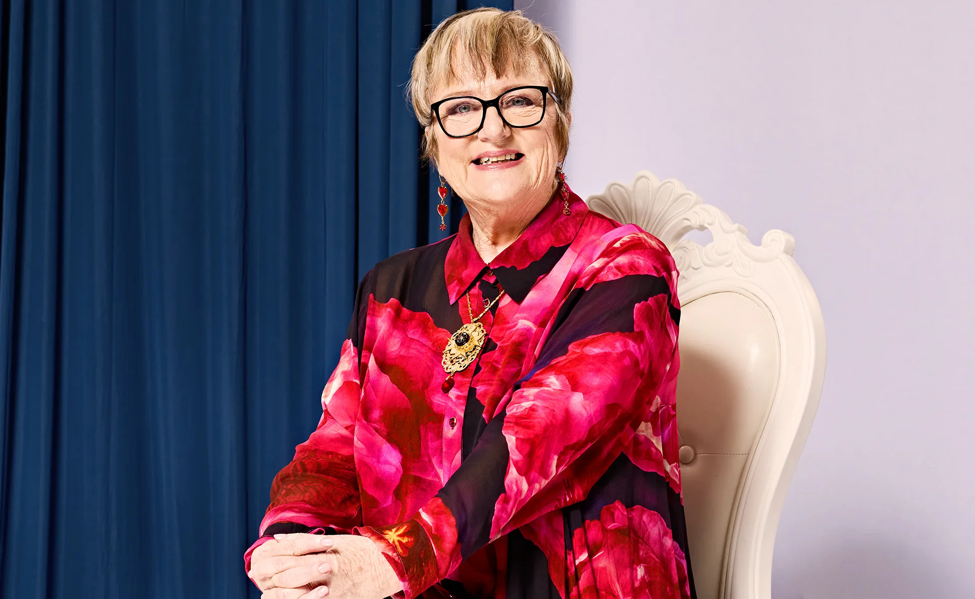 Denise, wearing a red and black jacket and black-rimmed glasses, sits in a chair