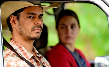 Mark Coles Smith, wearing a cowboy hat, sits in the front seat of a car, next to Tuuli Narkle, on the set of Mystery Road: Origin