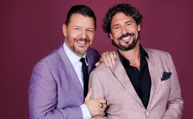 Manu and Colin dressed in nice suits, smiling against a maroon backdrop for My Kitchen Rules 2025.