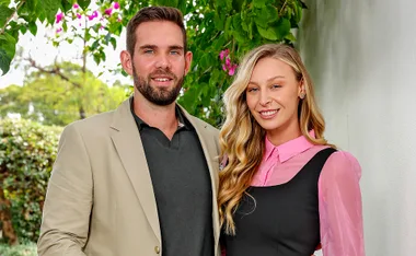 Michael, in a black shirt and beige jacket, and Rielli, in a pink and black outfit, stand together on the street outside a home