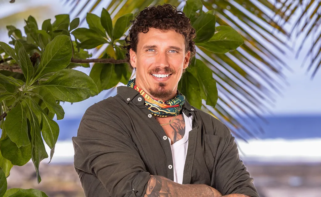 Luke stands on the beach in Samoa with a Survivor buff around his neck, smiling.