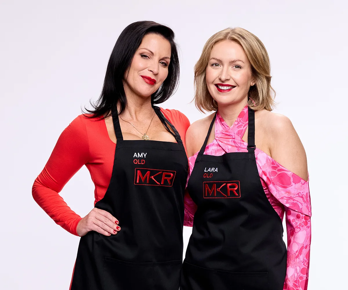 Amy and Lara, both wearing the black MKR aprons, stand with their arms around each other 