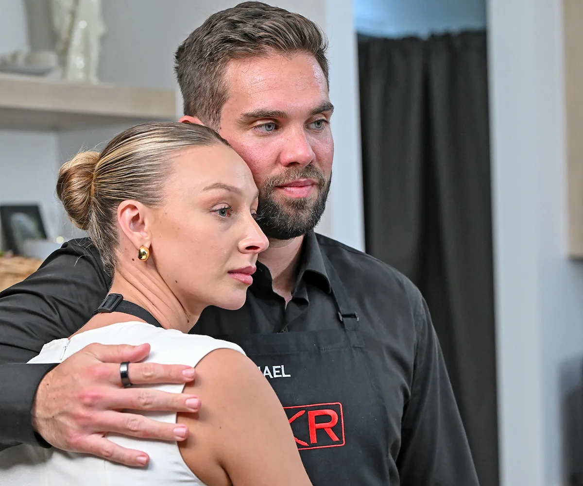 Michael, wearing a black jacket, puts his arm around Rielli, wearing a white dress,  while the two stand in their kitchen