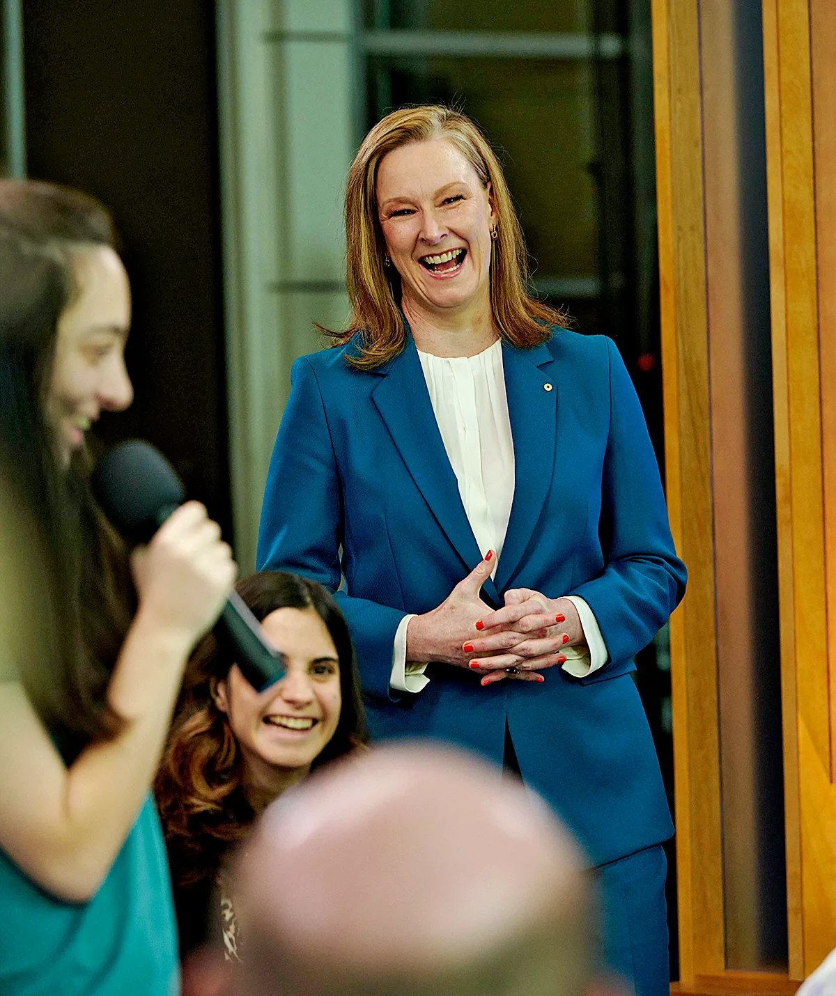 Leigh, wearing a blue jacket, smiles as one of the students holds a microphone 