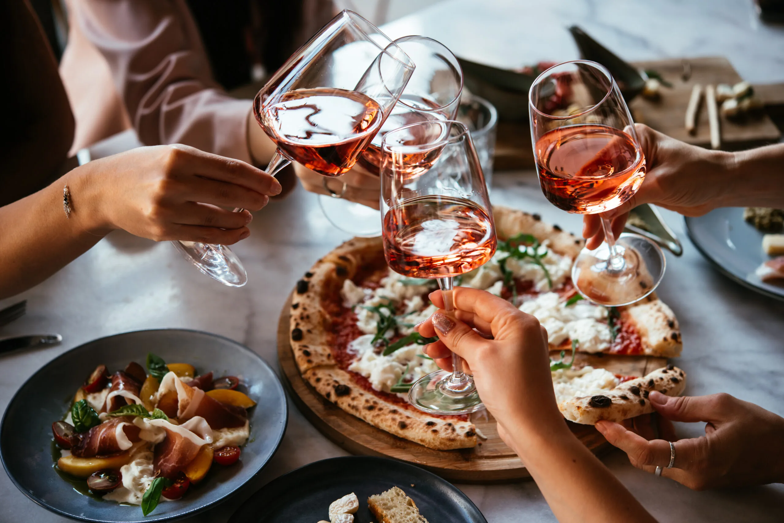 Friends clink wine glasses over a pizza.