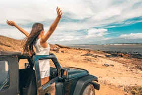 A woman jubilantly throws her arms in the air as she stands in an open-top car on a beach.