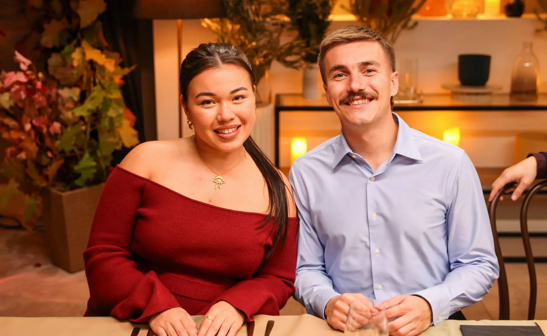 Danielle, in a red dress, and Marko, in a pale shirt, sit next to each other at a dinner table