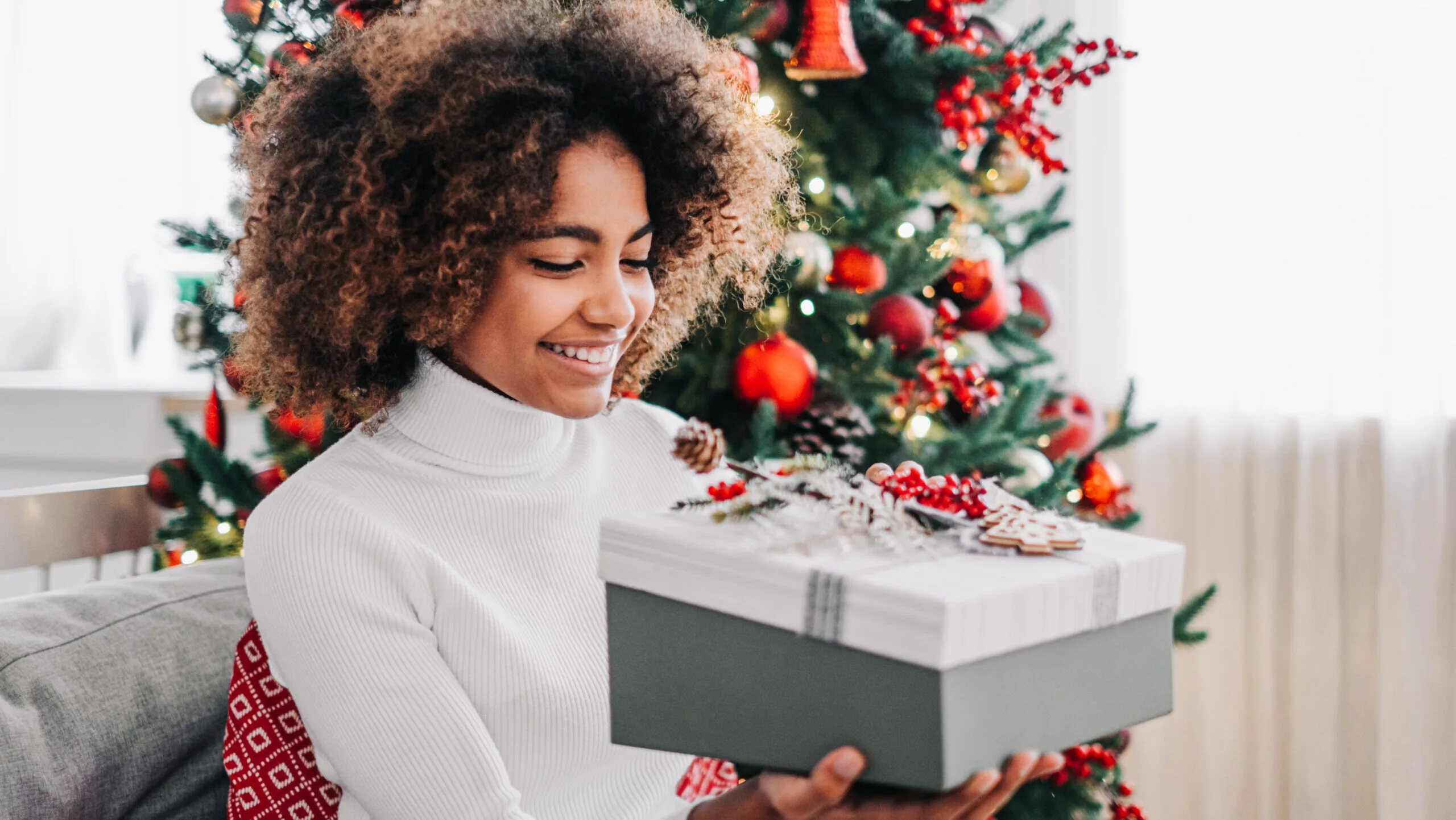 young girl opens a Christmas gift at home on the sofa against the background of a festive Christmas tree.