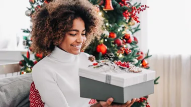 young girl opens a Christmas gift at home on the sofa against the background of a festive Christmas tree.