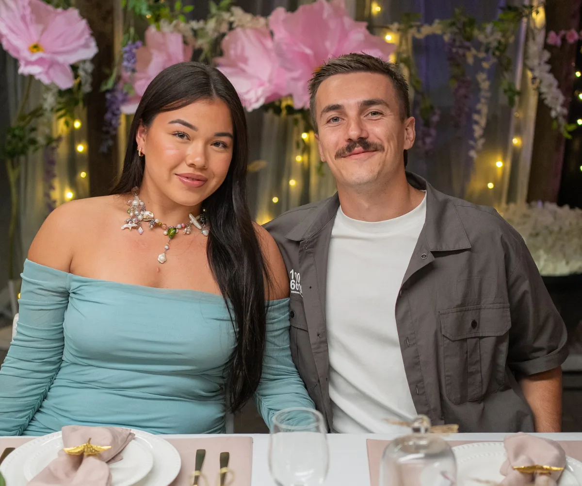 Danielle, wearing a blue dress, sits next to Marko, wearing a grey shirt over a white t-shirt, at a dinner table