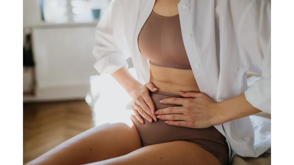 A woman in tan brown underwear with a white overshirt clutches her stomach.