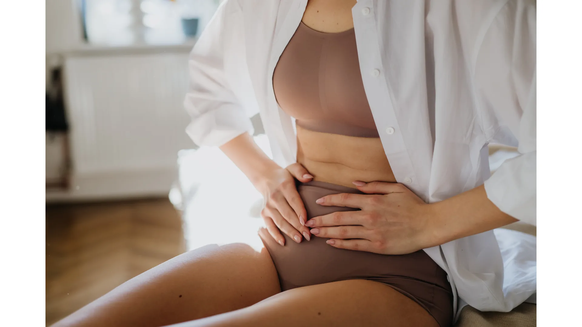 A woman in tan brown underwear with a white overshirt clutches her stomach.