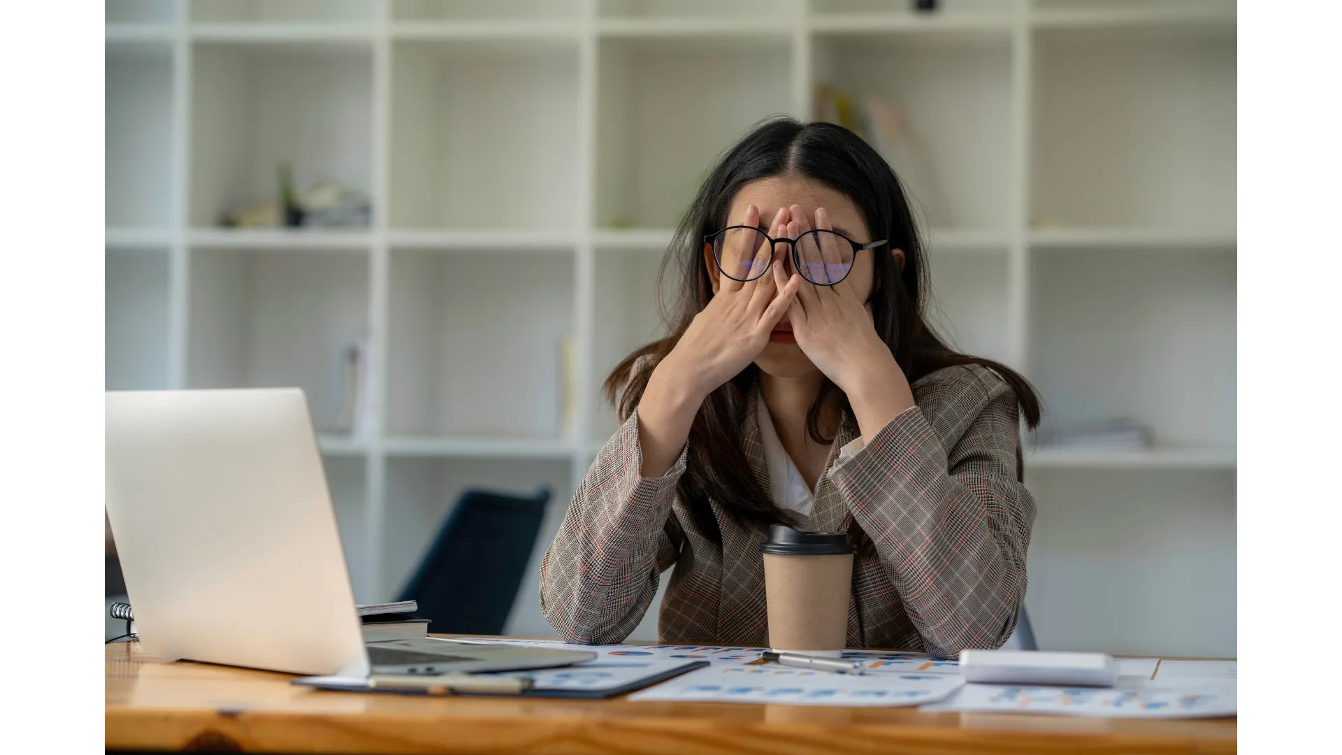 A stressed and exhausted woman rubs her eyes underneath her glasses.