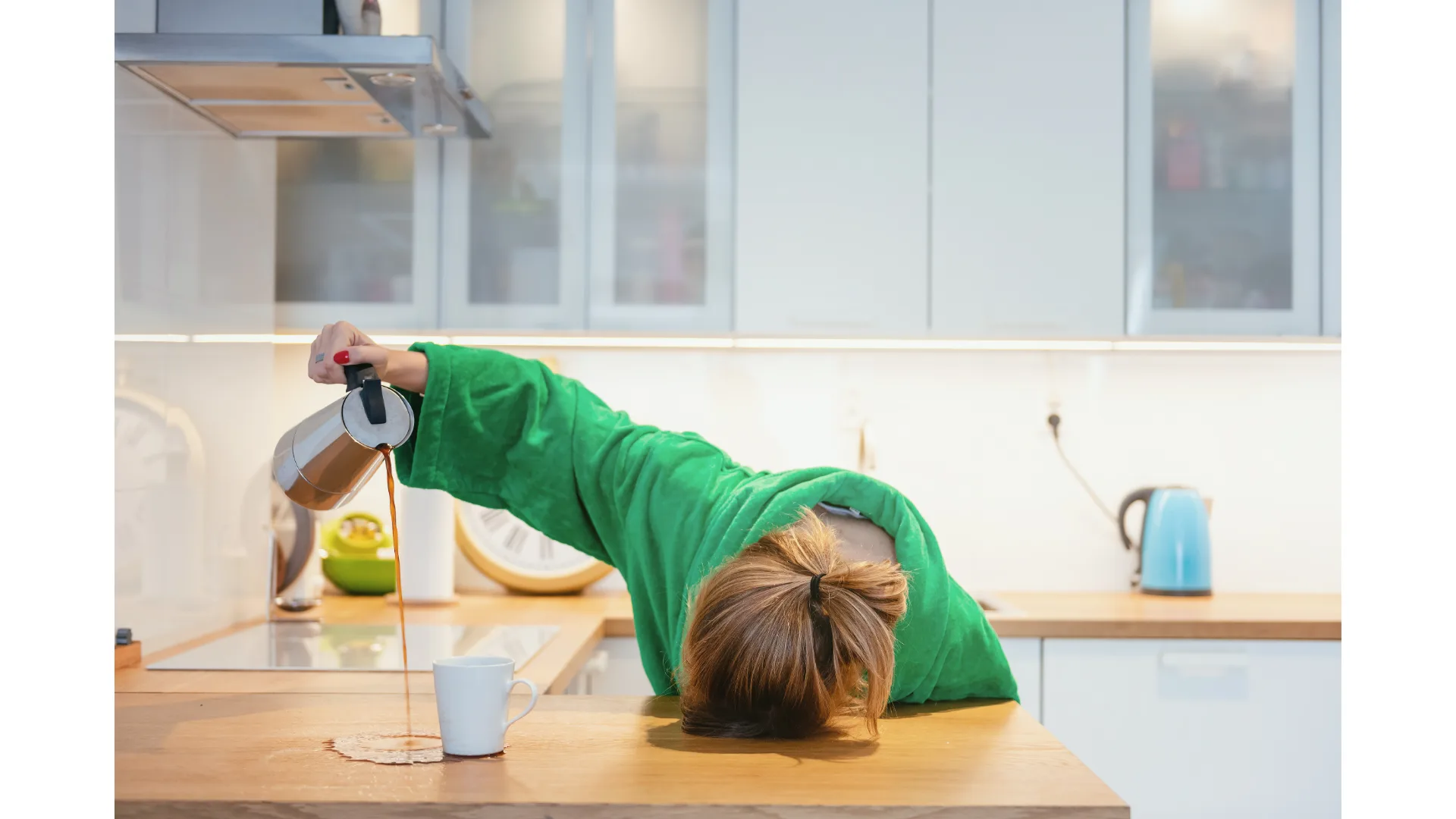 An exhausted woman slumps over her kitchen counter while pouring coffee onto the bench instead of into her mug. 