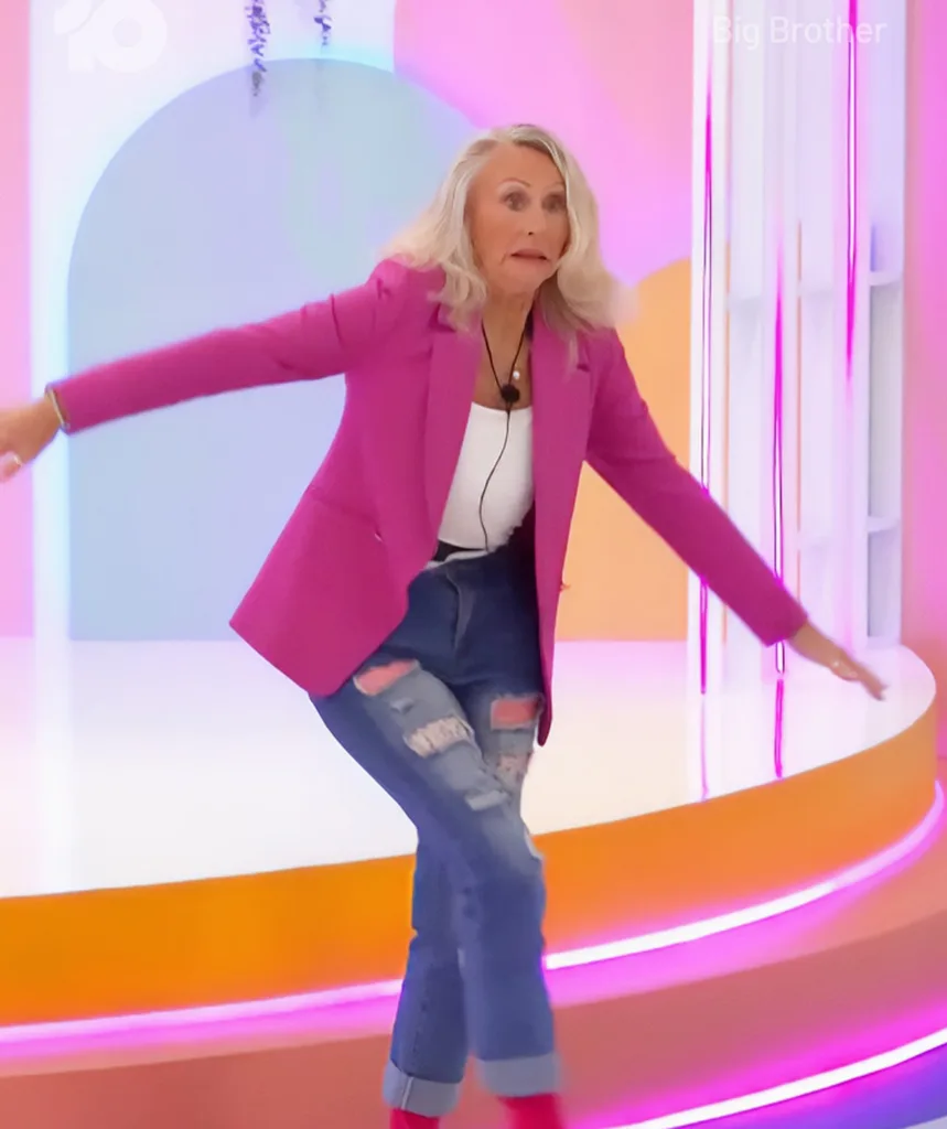 Jane, wearing a pink jacket and ripped jeans, stands on one leg, looking awkward