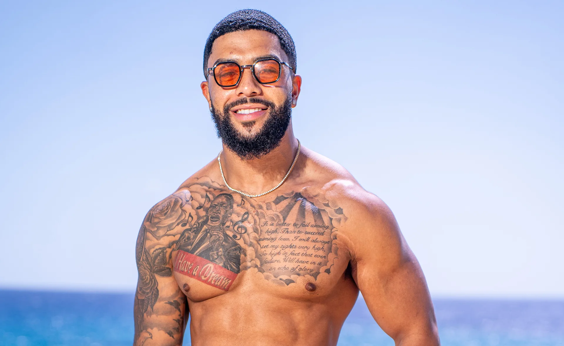 Dylan shirtless on a beach with sunglasses smiling on Love Island Australia 2025.