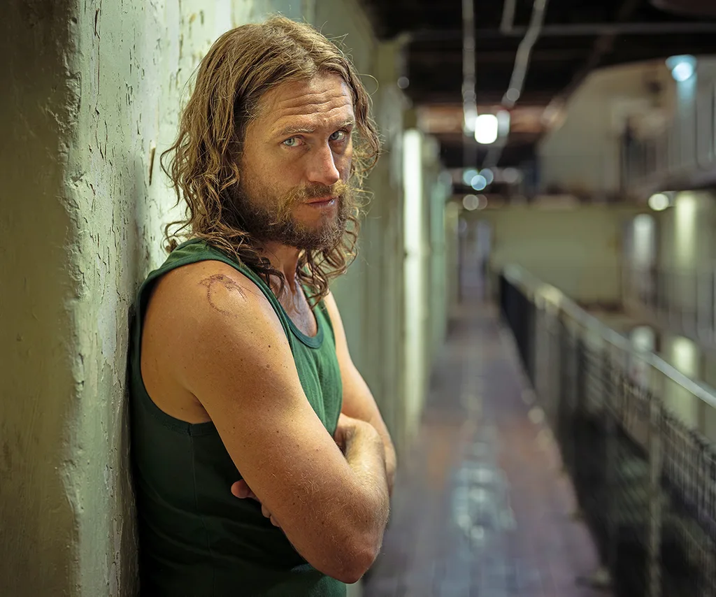 George, in character as Brenden, wears a green singlet and stands with his arms folded inside a prison during the filming of drama series Run