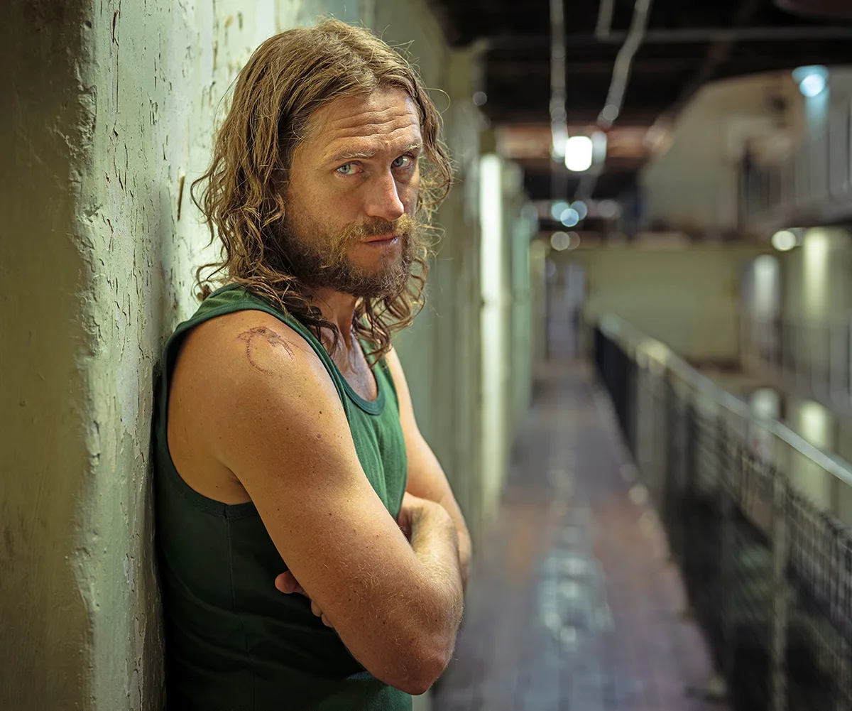 George, in character as Brenden, wears a green singlet and stands with his arms folded inside a prison during the filming of drama series Run