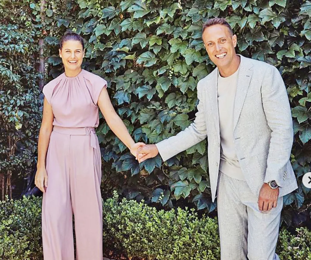 Matt, in a pale grey suit, holds hands with his wife Jessica, in a pale pink suit, in front of a hedge.