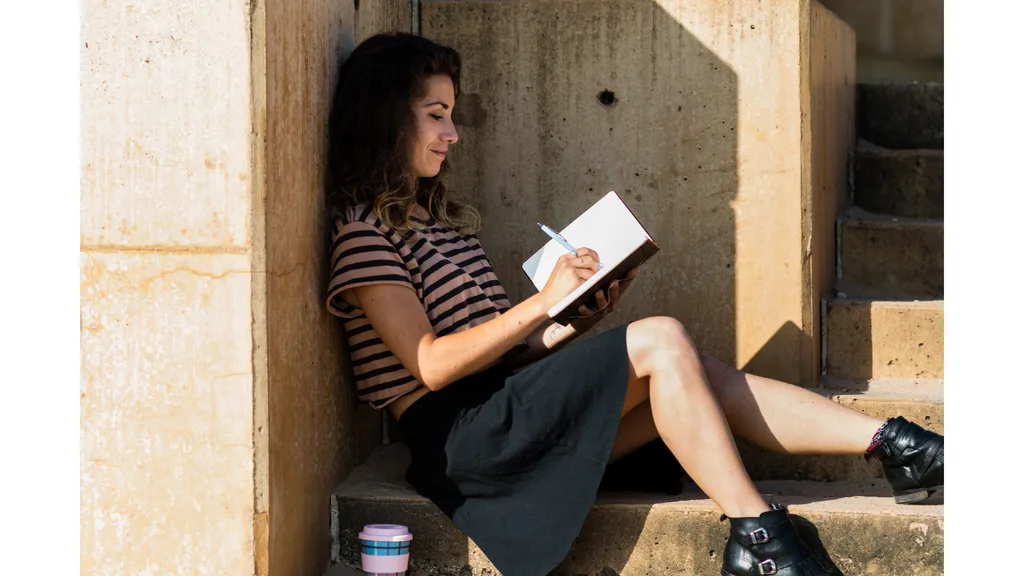 A young woman sits outside on a step while writing in a journal.