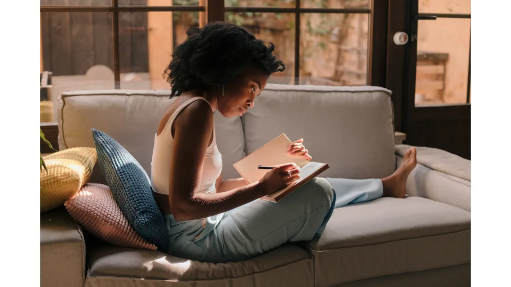 A young woman sits on a couch while writing in a diary.