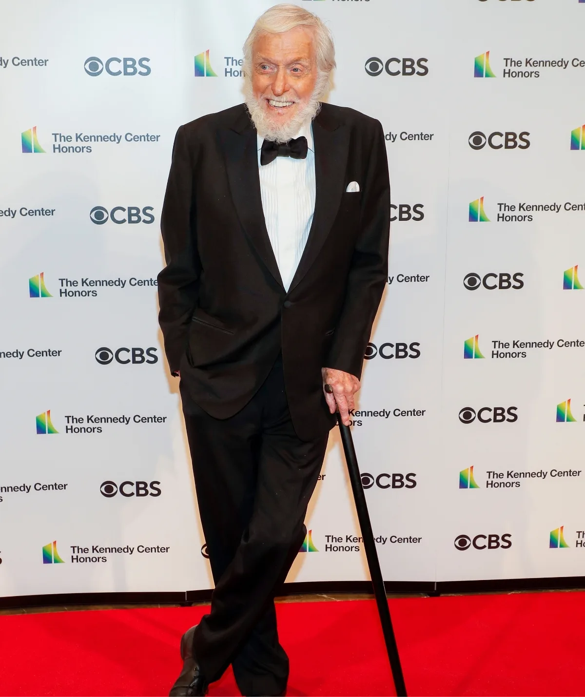 An older man in a tuxedo on a red carpet, leaning on a walking stick.
