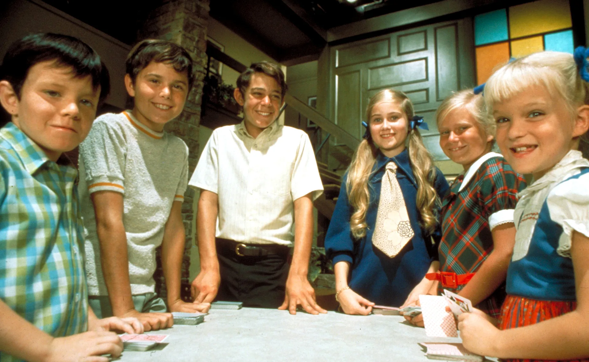 The six child actors who played the Brady Bunch kids stand around a table in a dark room