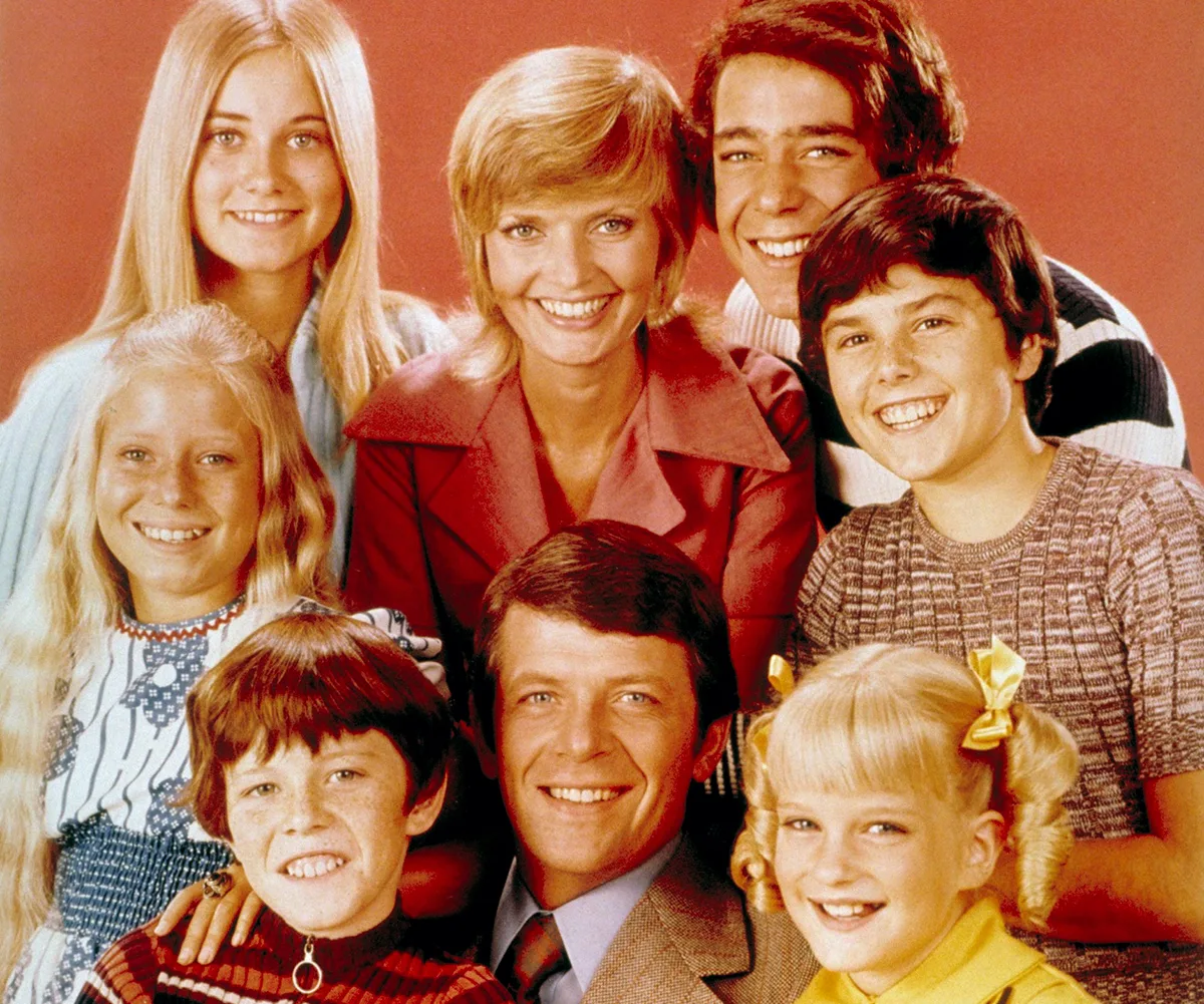 Florence Henderson, as Carol, and Robert Reed, as Mike, are surrounded by the six actors playing their onscreen kids, in front of a brown background