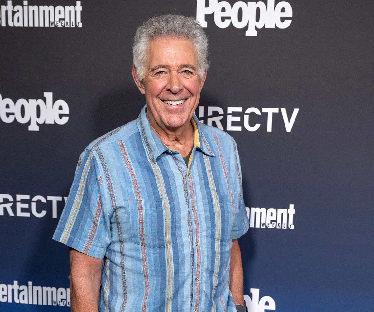 Barry Williams, with greying hair, wearing a striped shirt, stands in front of a background reading DIRECTV and People