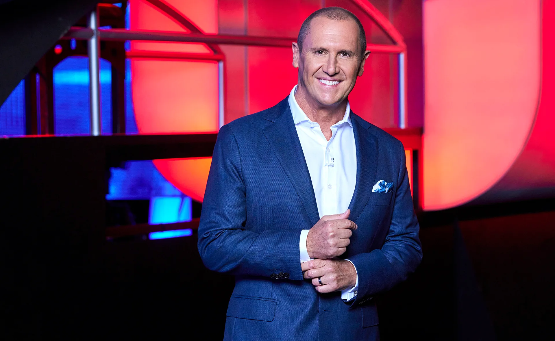 Larry Emdur, wearing a white shirt and blue suit, stands on the set of The Chase