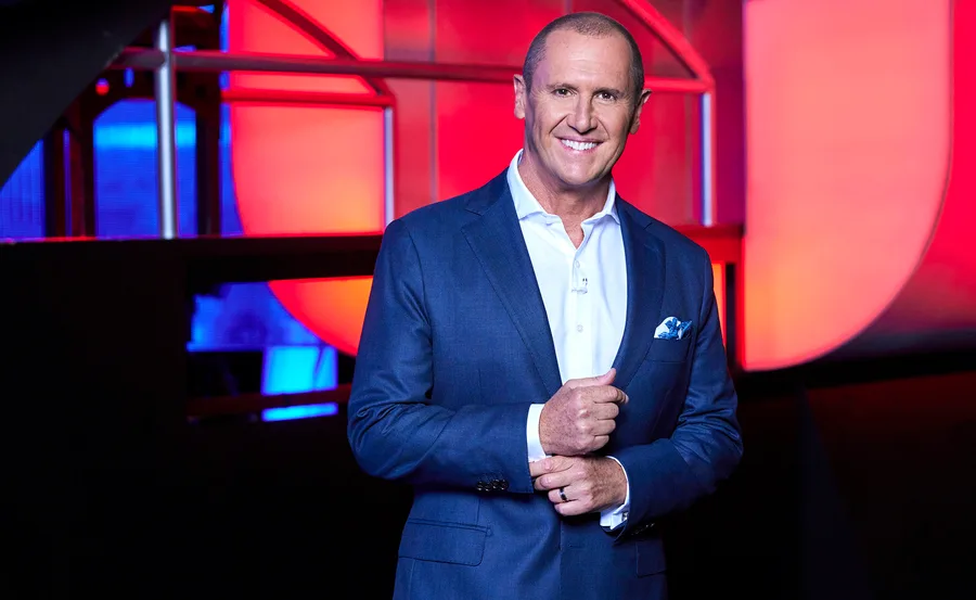 Larry Emdur, wearing a white shirt and blue suit, stands on the set of The Chase